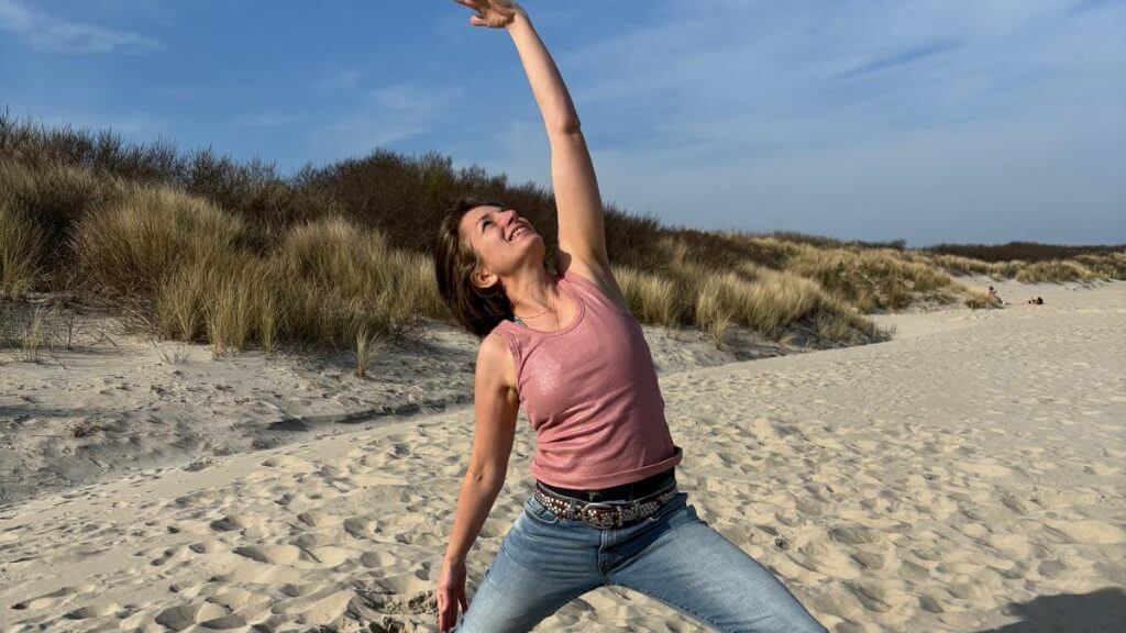Frau in Jeans streckt in einer Yoga-Pose einen Arm zum Himmel, im Hintergrund Dünen und Strand.
