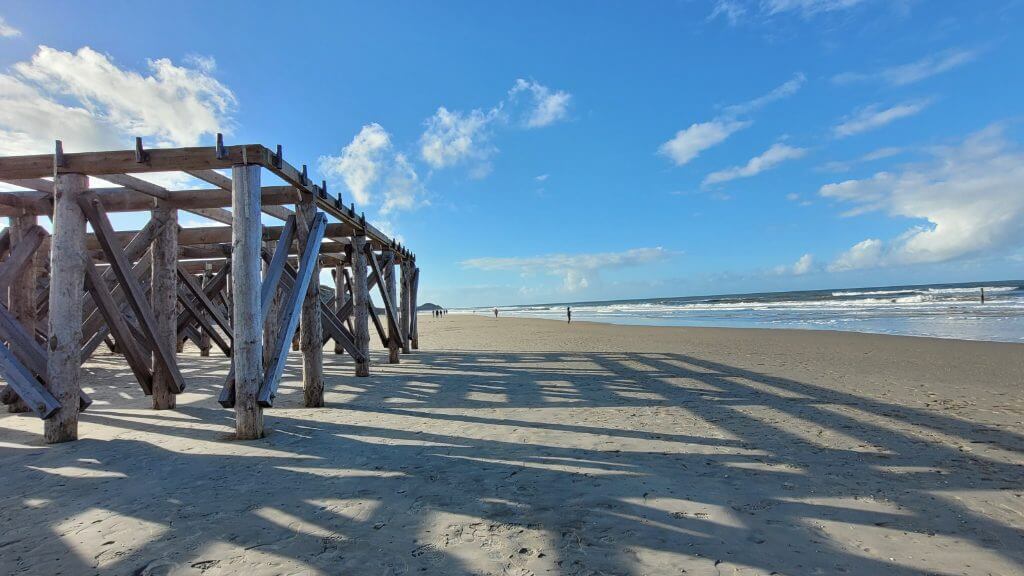 Ein Holzsteg wirft lange Schatten auf den Sandstrand unter einem teilweise bewölkten Himmel.