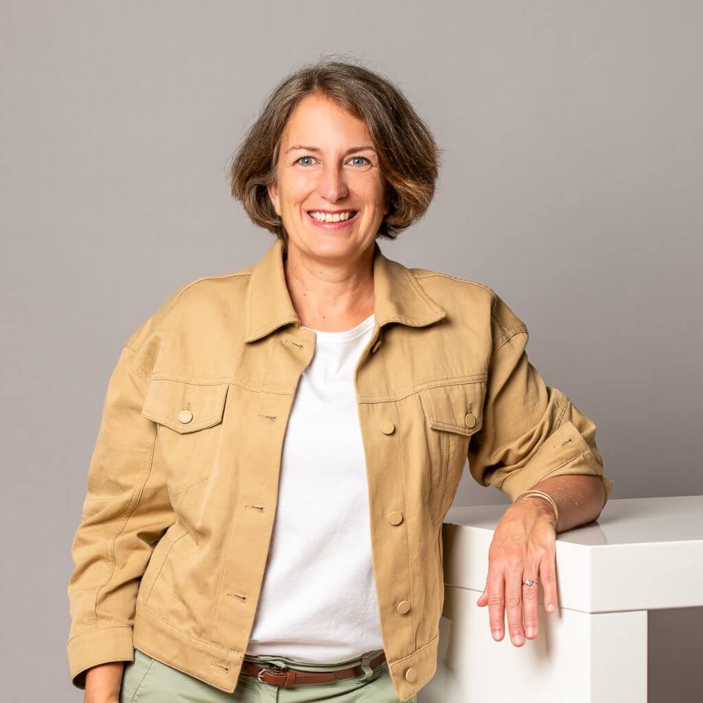 Nahaufnahme von Sandra Krönner mit freundlichem Lächeln und beige Jacke vor neutralem Hintergrund.