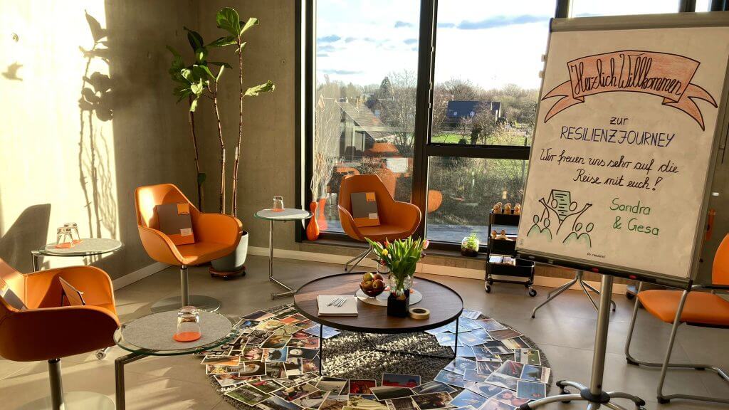 Heller Seminarraum mit orangen Sesseln, ausgelegten Bildkarten und einem Flipchart zur Resilienz-Journey.