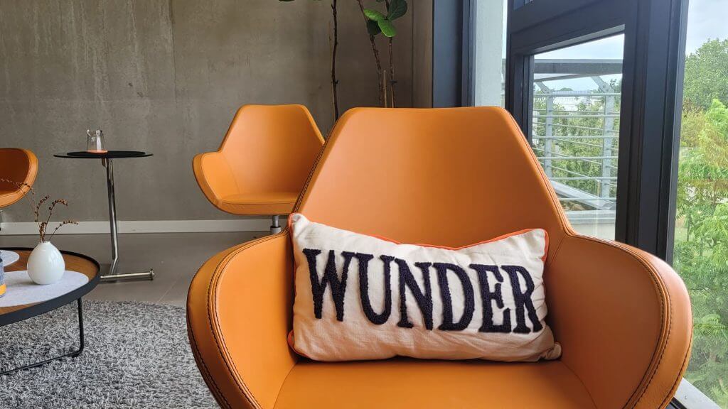 Orangefarbener Sessel mit einem Kissen mit der Aufschrift WUNDER in einem hellen Seminarraum mit großer Fensterfront.