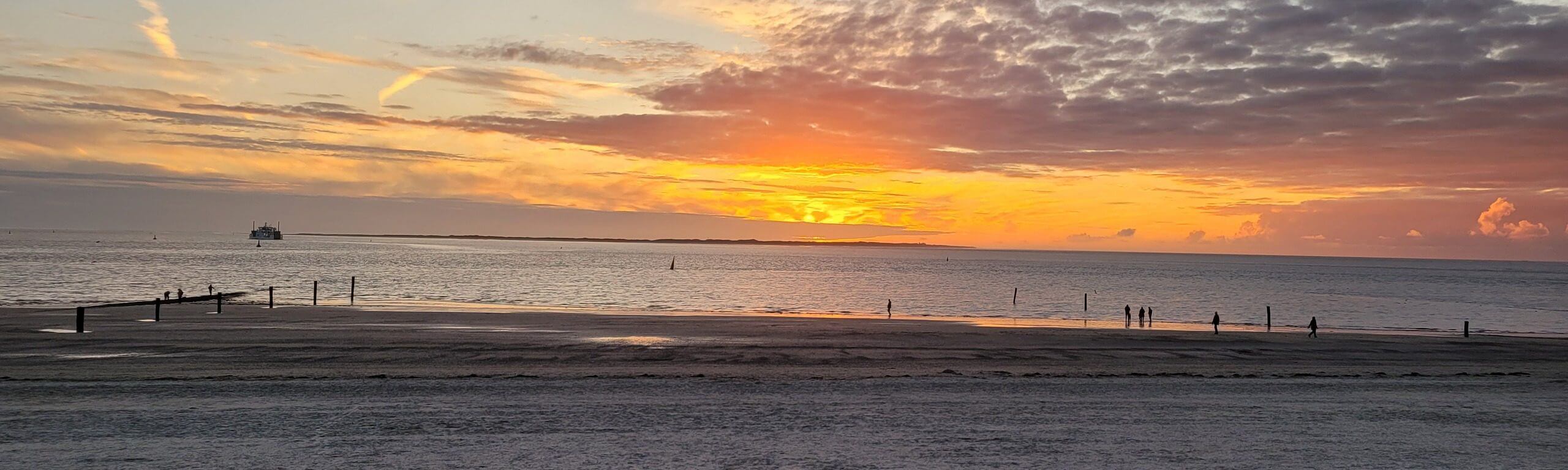 Sonnenuntergang über dem Meer, am Strand gehen vereinzelt Menschen spazieren.