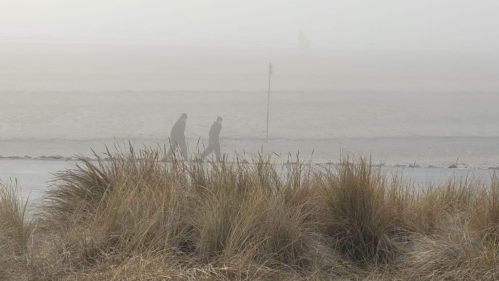 Zwei Personen gehen an einem nebligen Strand entlang, umgeben von Dünen.