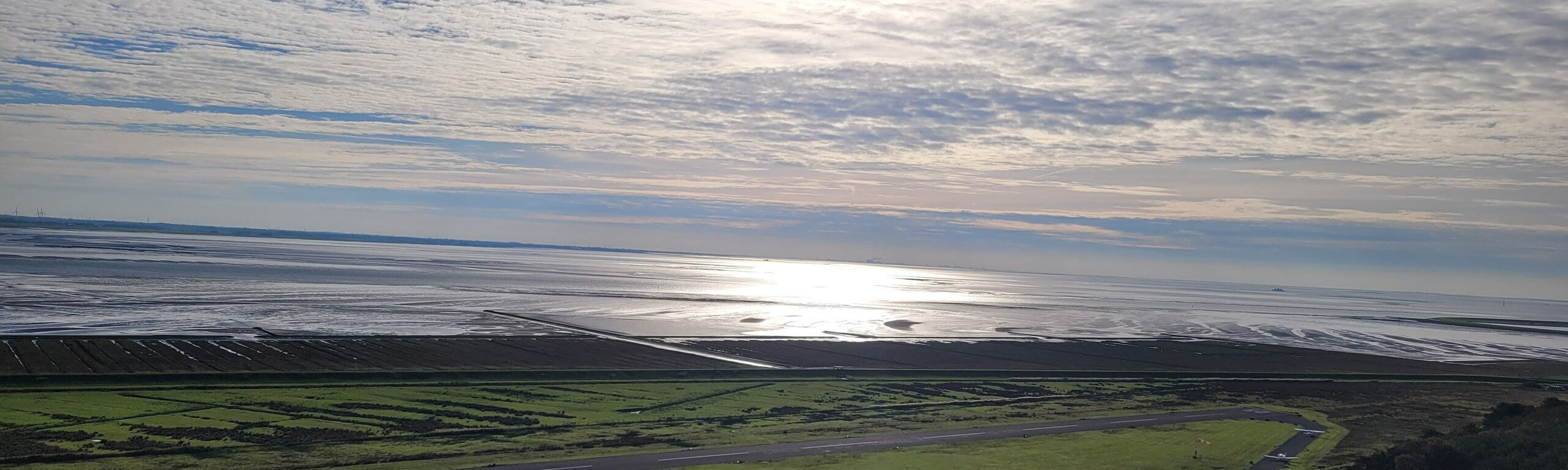Blick von oben auf Küstenwiesen und das Wattenmeer, auf dem sich das Licht unter Wolken spiegelt.