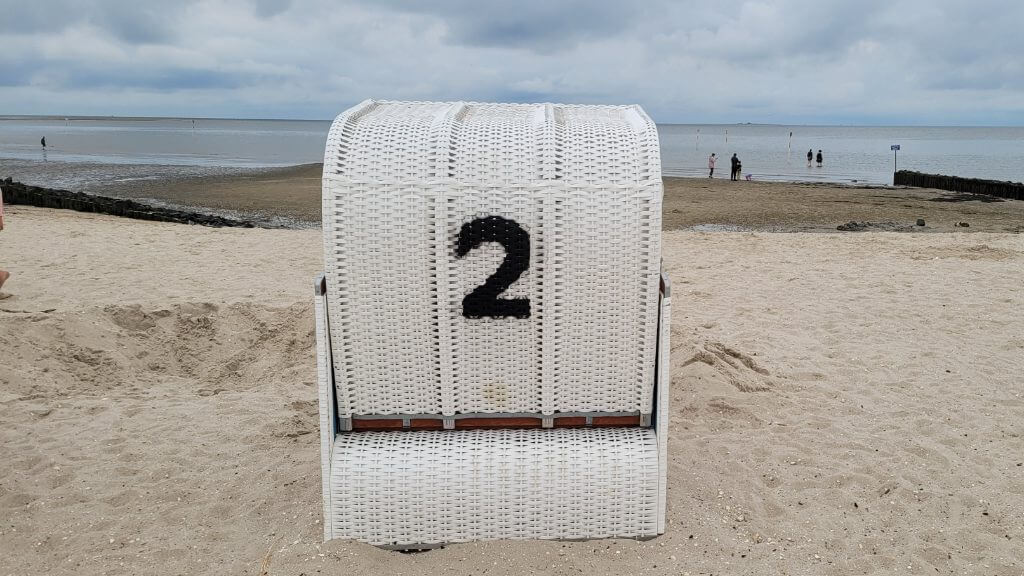 Ein weißer Strandkorb mit der Nummer 2 steht auf sandigem Boden am Meer.